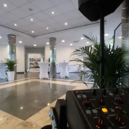 Sala konferencyjna przygotowana na wydarzenie, widoczne stoły koktajlowe w białych pokrowcach, kolumny, rośliny doniczkowe i sprzęt nagłośnieniowy. Mikser dźwięku na pierwszym planie.