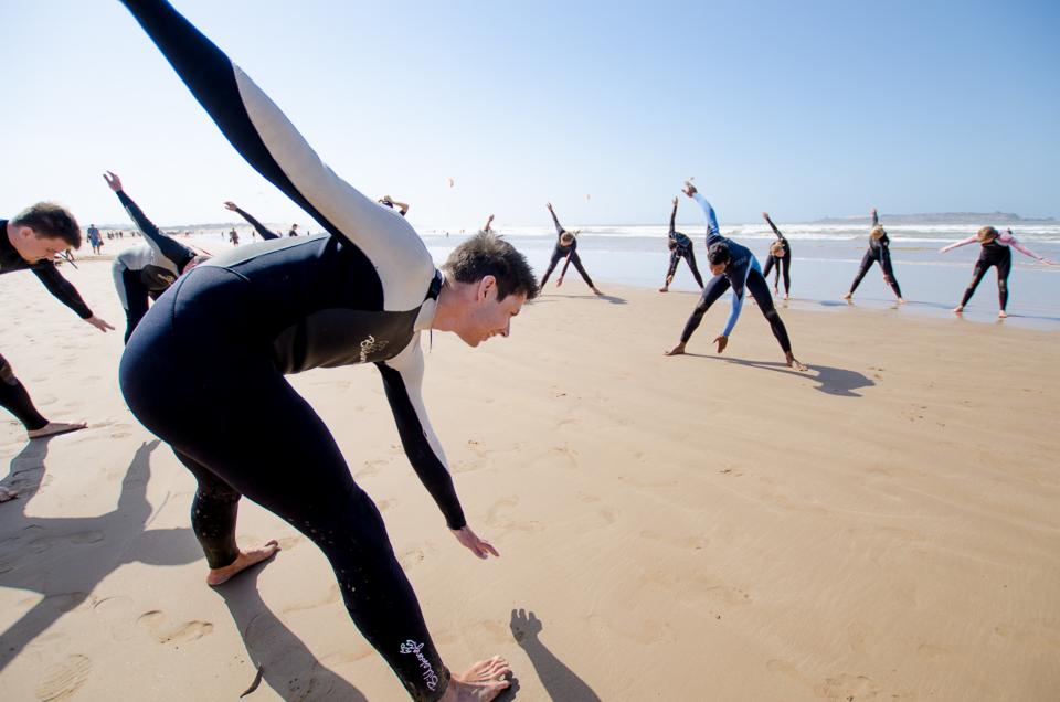 Grupa osób w piankach rozciąga się na plaży w słoneczny dzień, przygotowując się do aktywności wodnych; widoczne morze w tle.