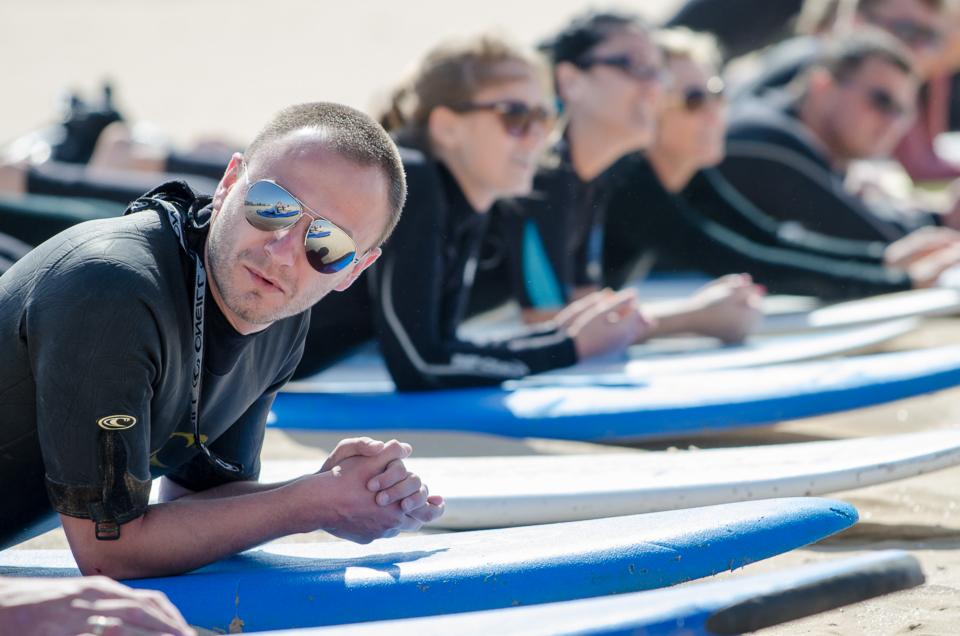 Grupa osób w czarnych piankach leżących na deskach surfingowych na plaży, odbicie desek i nieba w okularach mężczyzny z przodu.