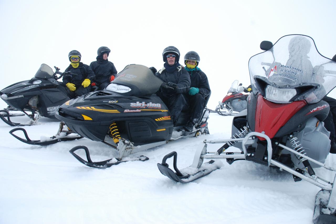 Grupa osób w kaskach i zimowych ubraniach siedzi na skuterach śnieżnych Ski-Doo i Yamaha Venture w zasypanym śniegiem terenie. Na przedniej szybie skutera widoczna naklejka 'Mountaineers of Iceland...