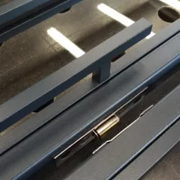 Elementy konstrukcyjne bramy wjazdowej z zamkiem, malowane proszkowo na szaro, leżące na stole warsztatowym.