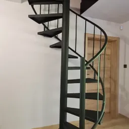 Czarna, metalowa schody spiralne w jasnym wnętrzu, widok z dołu, z drewnianą podłogą i drzwiami w tle.