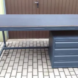 Metalowy stół warsztatowy z szufladami w kolorze grafitowym, na zewnątrz, z widoczną konstrukcją spawaną i drewnianym blatem.