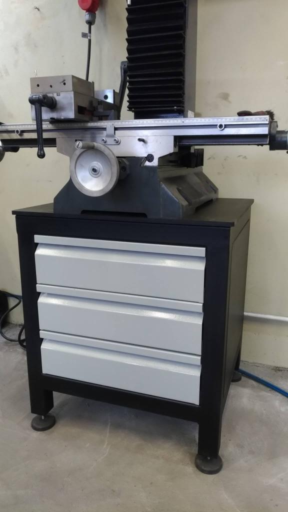 Metalowa frezarka warsztatowa z trzema szufladami na narzędzia, ustawiona na betonowej posadzce, z widocznymi elementami mocującymi i regulacyjnymi.