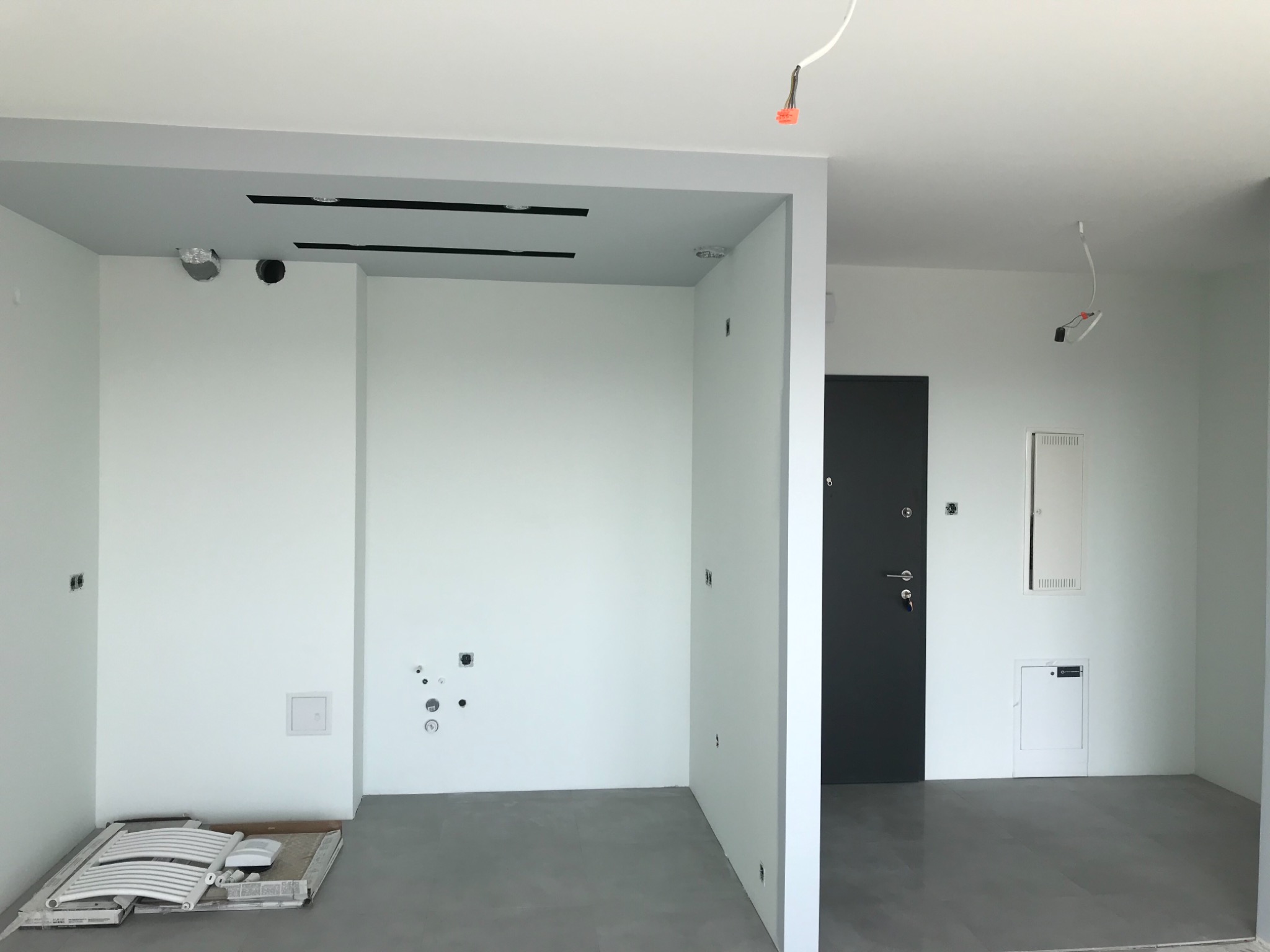 Wnętrze mieszkania w stanie deweloperskim z przygotowanymi otworami na instalacje, szarą podłogą i białymi ścianami, widoczne niedokończone podłączenia elektryczne.