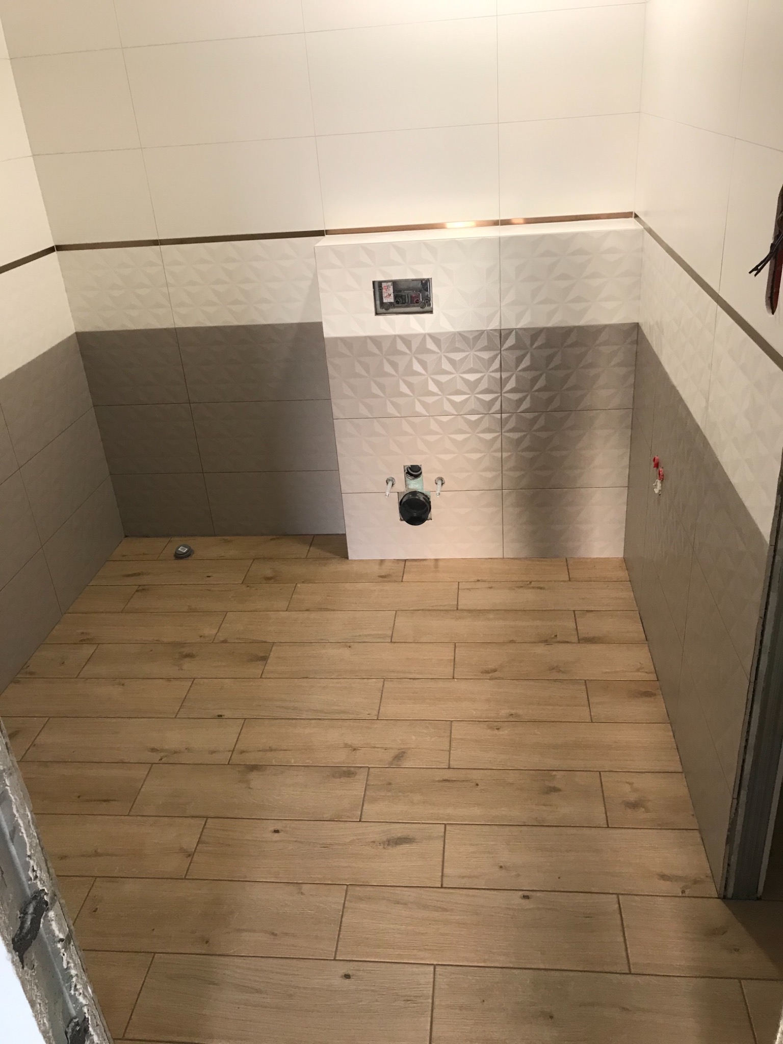 Wnętrze łazienki w trakcie remontu, widoczne płytki ścienne w odcieniach szarości i bieli z dekoracyjnym wzorem 3D, drewnopodobne płytki podłogowe, instalacja podtynkowa WC oraz fragment ościeżnicy...