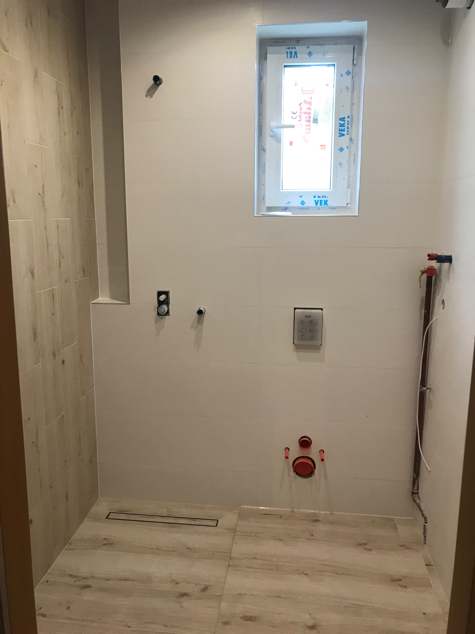 Wnętrze łazienki w trakcie prac wykończeniowych, widoczne płytki imitujące drewno na podłodze i częściowo na ścianie, białe płytki na pozostałych ścianach, okno PCV z folią ochronną, instalacja...