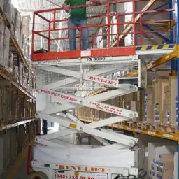 Pracownik w pomarańczowym kasku na podnośniku nożycowym marki Rent-Lift, wykonujący prace na wysokości w magazynie z regałami