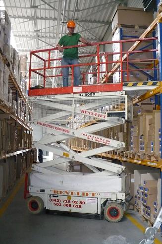 Pracownik w pomarańczowym kasku na podnośniku nożycowym marki Rent-Lift, wykonujący prace na wysokości w magazynie z regałami