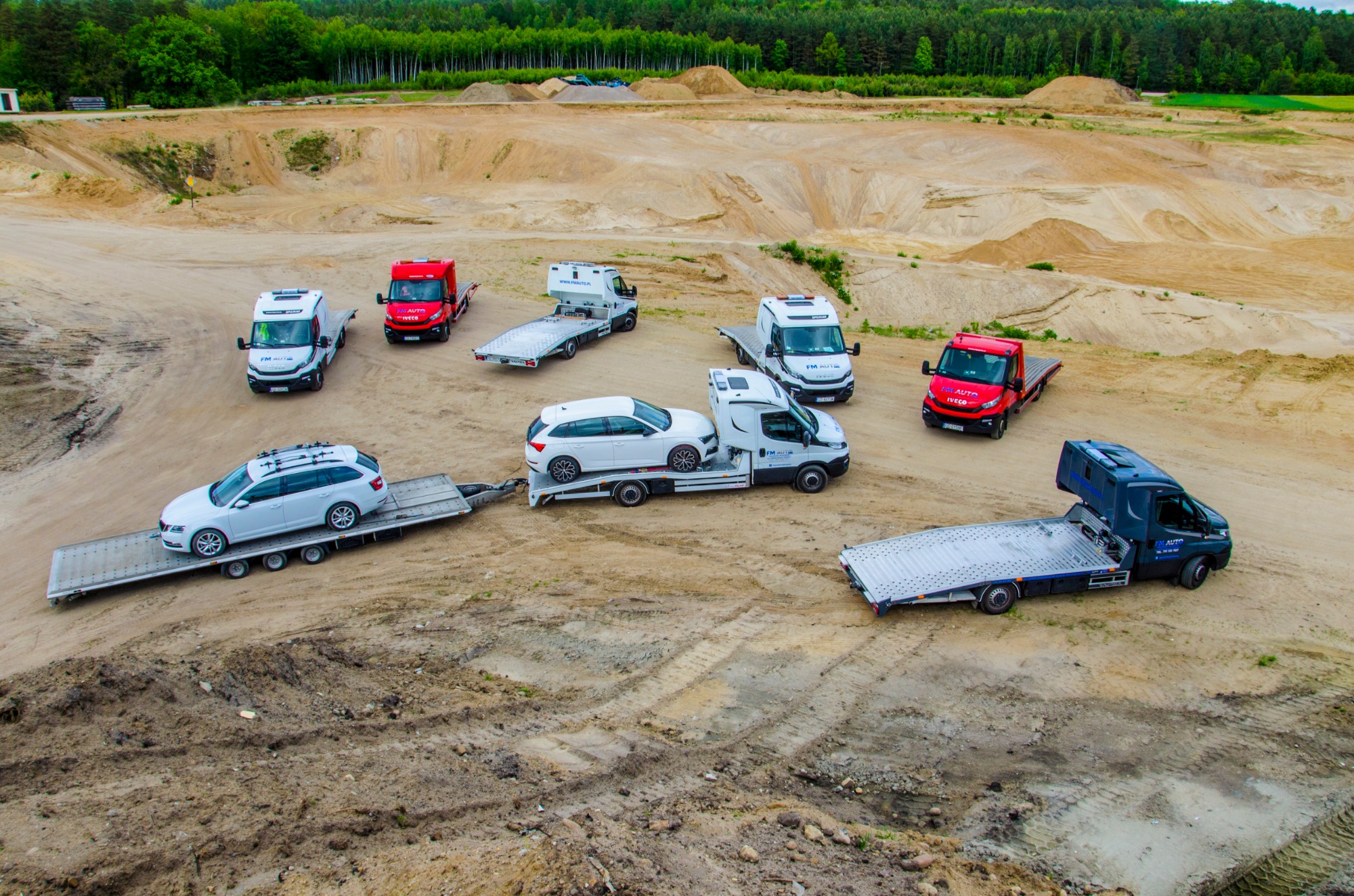 Widok z góry na flotę lawet i przyczep z samochodami na piaszczystym placu budowy; w tle las.