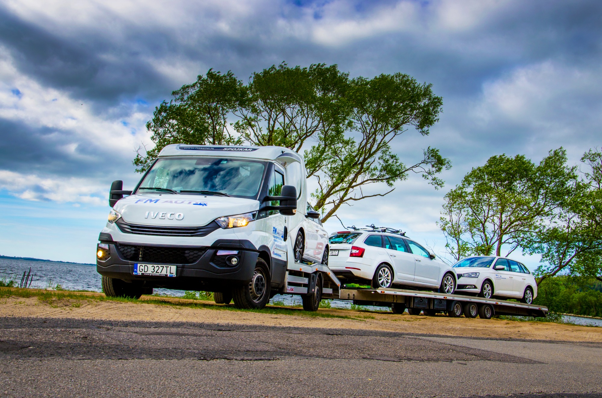 Biała laweta Iveco z trzema białymi samochodami Skoda na platformie, zaparkowana na poboczu drogi z widokiem na jezioro i drzewa w tle pod pochmurnym niebem.