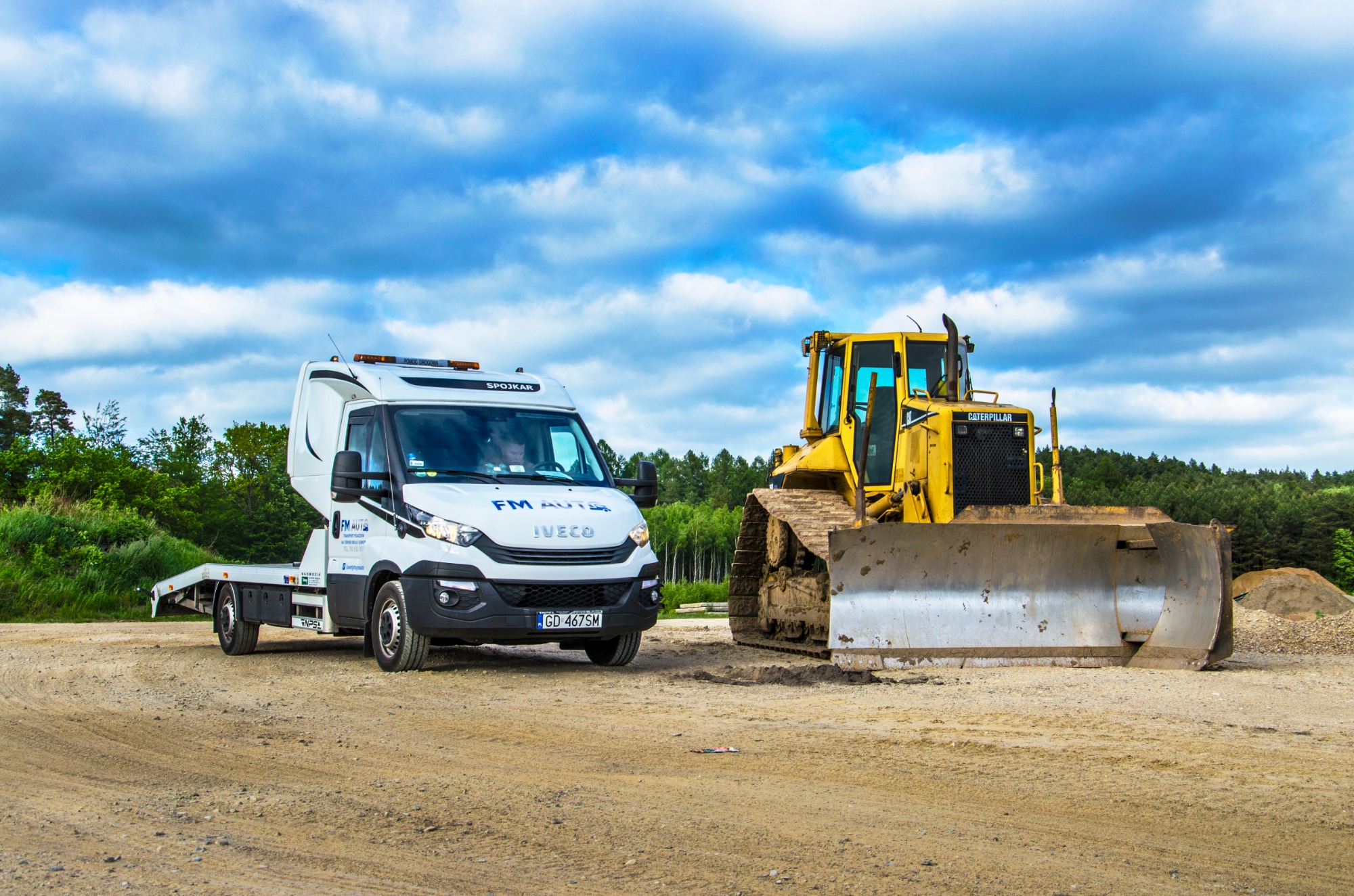 Biała laweta Iveco z pomarańczowymi światłami ostrzegawczymi i logo FM Auto, obok żółtej spycharki Caterpillar na piaszczystym placu budowy z widokiem na las i błękitne niebo z chmurami.