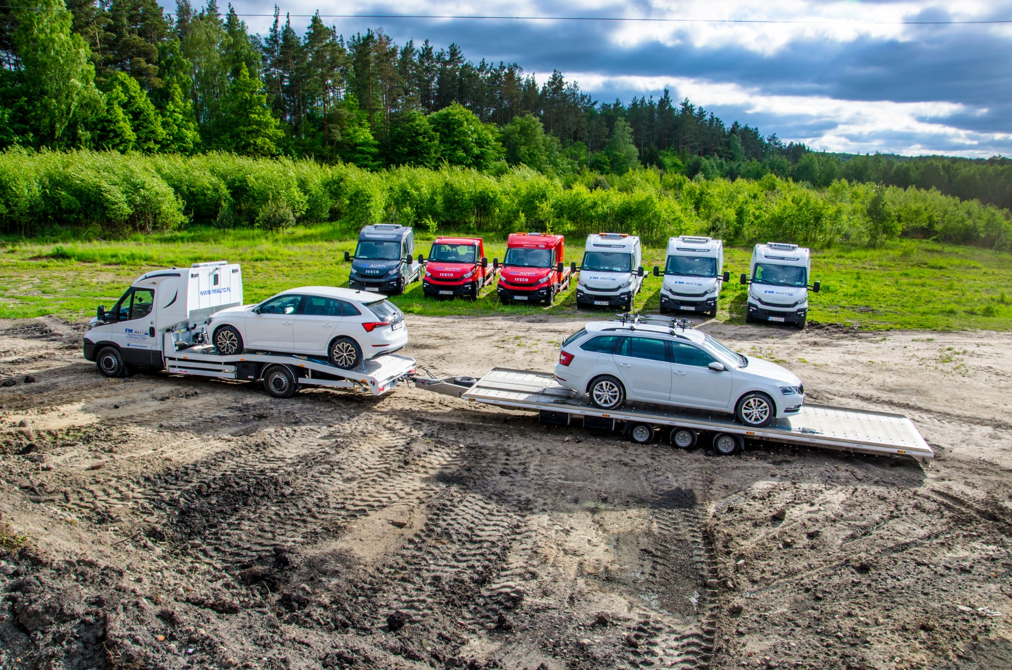 Dwa białe samochody typu kombi na lawetach, w tle rząd zaparkowanych busów na tle lasu, teren błotnisty z widocznymi śladami opon.