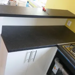 Czarny, nakrapiany blat kuchenny z białymi szafkami i płytą gazową; na blacie leży czerwony śrubokręt, a na piekarniku naklejka z klasą energetyczną.