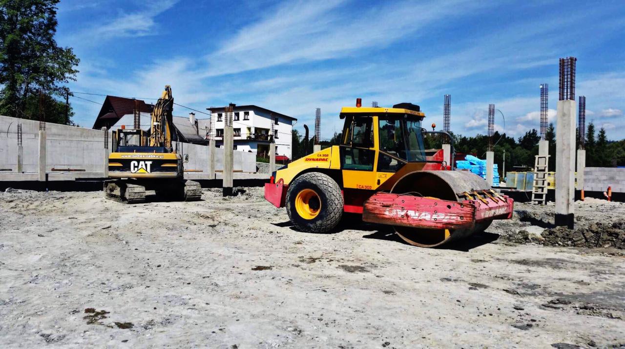 Żółto-czerwony walec drogowy Dynapac CA302 i koparka CAT na placu budowy, widoczne fundamenty i zbrojenia, w tle budynki mieszkalne i błękitne niebo.