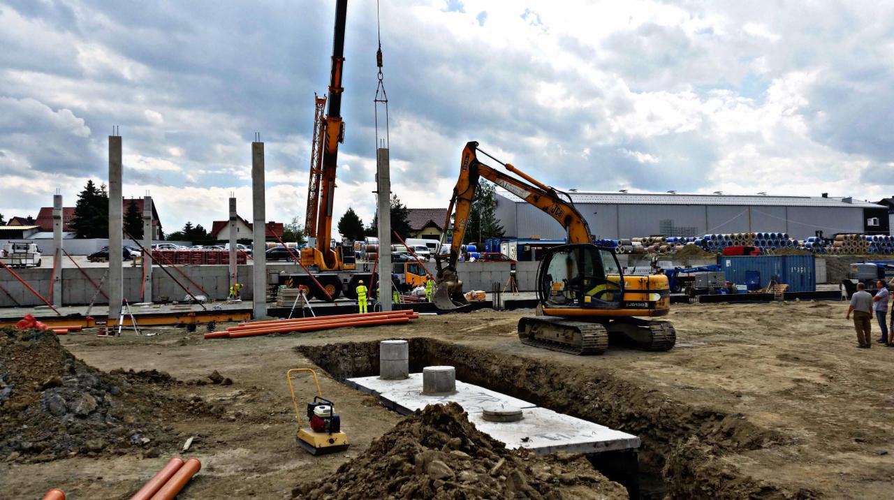 Plac budowy z dźwigiem podnoszącym betonowy słup, obok koparka JCB, ubijarka gruntu i fundamenty pod budynek przemysłowy w tle.