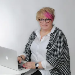 Kobieta w okularach i marynarce w pepitkę, z różowymi pasemkami, siedzi przy białym stoliku i pracuje na laptopie.