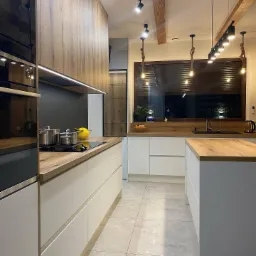 Nowoczesna kuchnia z białymi szafkami, drewnianym blatem i wyspą, oświetlona lampami wiszącymi na sznurach, z widocznymi garnkami na płycie i drewnianymi belkami na suficie.