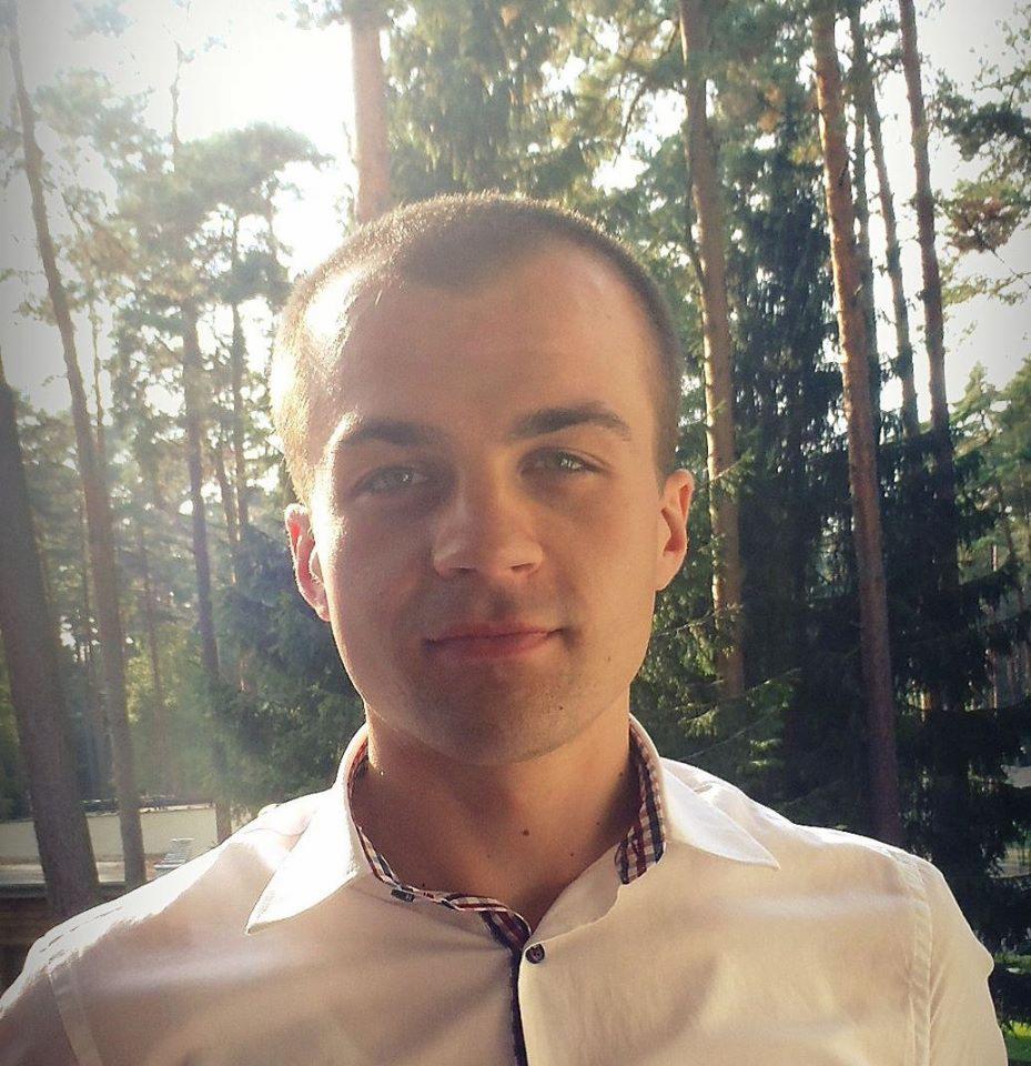 Portret młodego mężczyzny w białej koszuli z wzorzystym kołnierzykiem na tle lasu iglastego w słoneczny dzień.