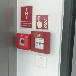 Czerwone przyciski alarmowe i tabliczki informacyjne zamontowane na białej ścianie wewnątrz budynku, widoczny przycisk POLAR i BOSCH.