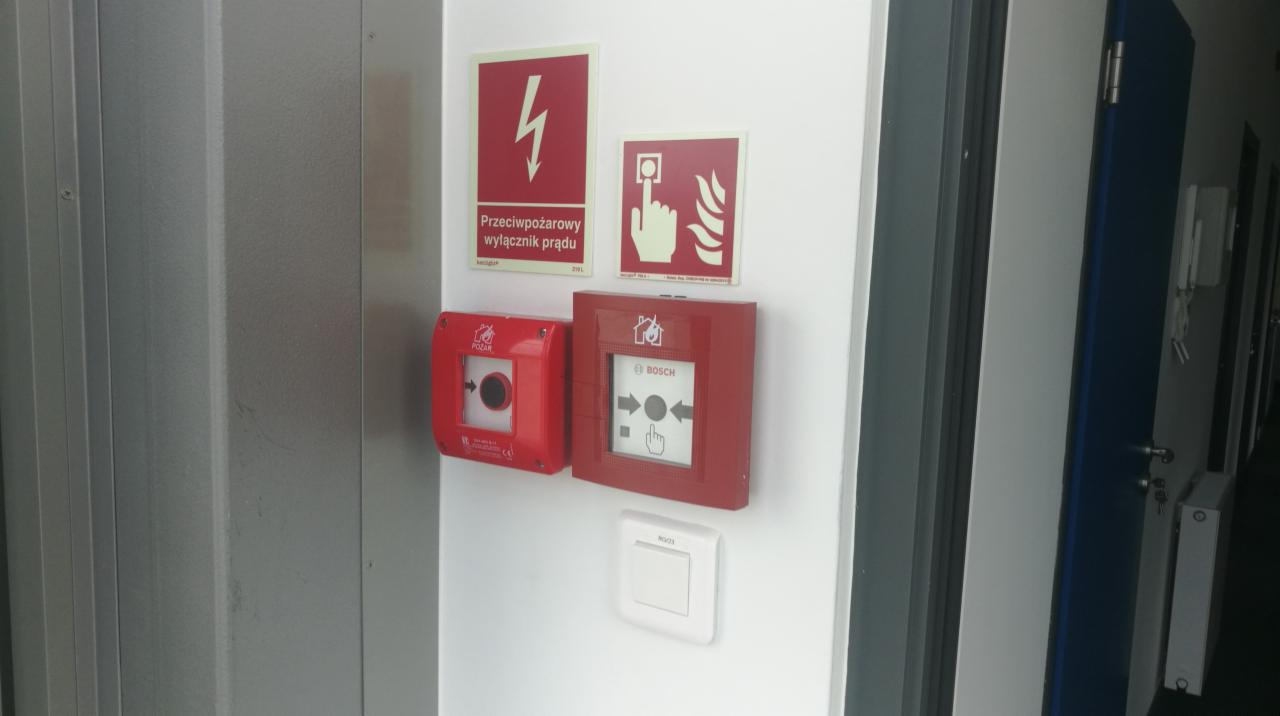 Czerwone przyciski alarmowe i tabliczki informacyjne zamontowane na białej ścianie wewnątrz budynku, widoczny przycisk POLAR i BOSCH.