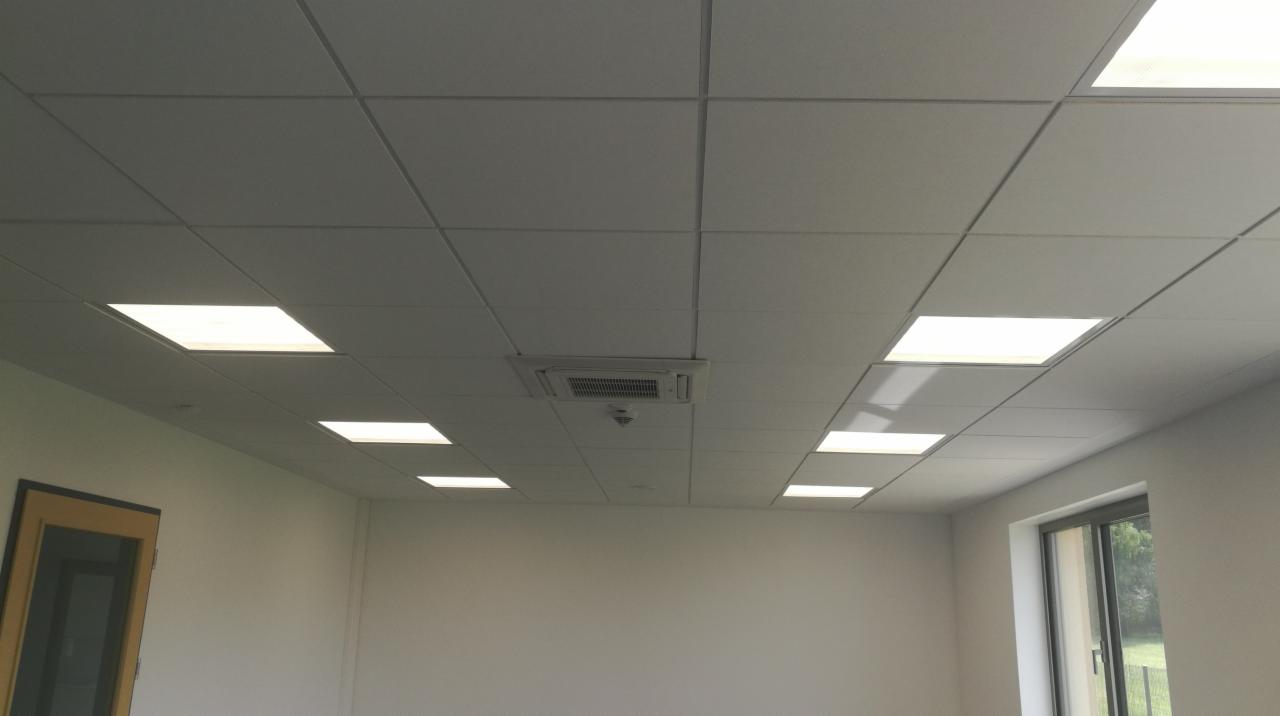 Kasetonowy sufit podwieszany z wbudowanymi panelami LED i centralną klimatyzacją, widok na fragment pomieszczenia z oknem i drzwiami.