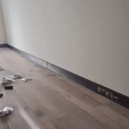 Montaż czarnych listew przypodłogowych w pomieszczeniu z drewnopodobną podłogą, widoczne narzędzia i akcesoria montażowe rozsypane na podłodze, ściany pomalowane na biało, okno w tle.