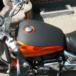 Motocykl z pomarańczowym zbiornikiem paliwa zdobionym płomieniami, czarnym skórzanym pokrowcem na zbiorniku i czarnym skórzanym siedzeniem; w tle stoi fotel.