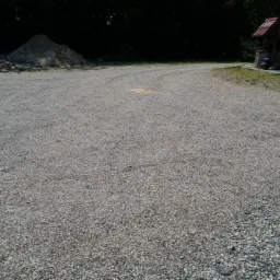 Utwardzony teren z szarego kruszywa, w tle koparka, pryzma ziemi i ozdobna studnia z roślinami.