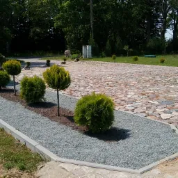 Ułożona z kamienia ścieżka prowadzi do studni, obok rabata z kulistymi krzewami i szarym żwirem, w tle zielony ogród i fragment budynku.