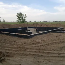 Zalane fundamenty domu jednorodzinnego z hydroizolacją, widoczne wystające pręty zbrojeniowe, teren wyrównany, w tle łąka i zabudowania.