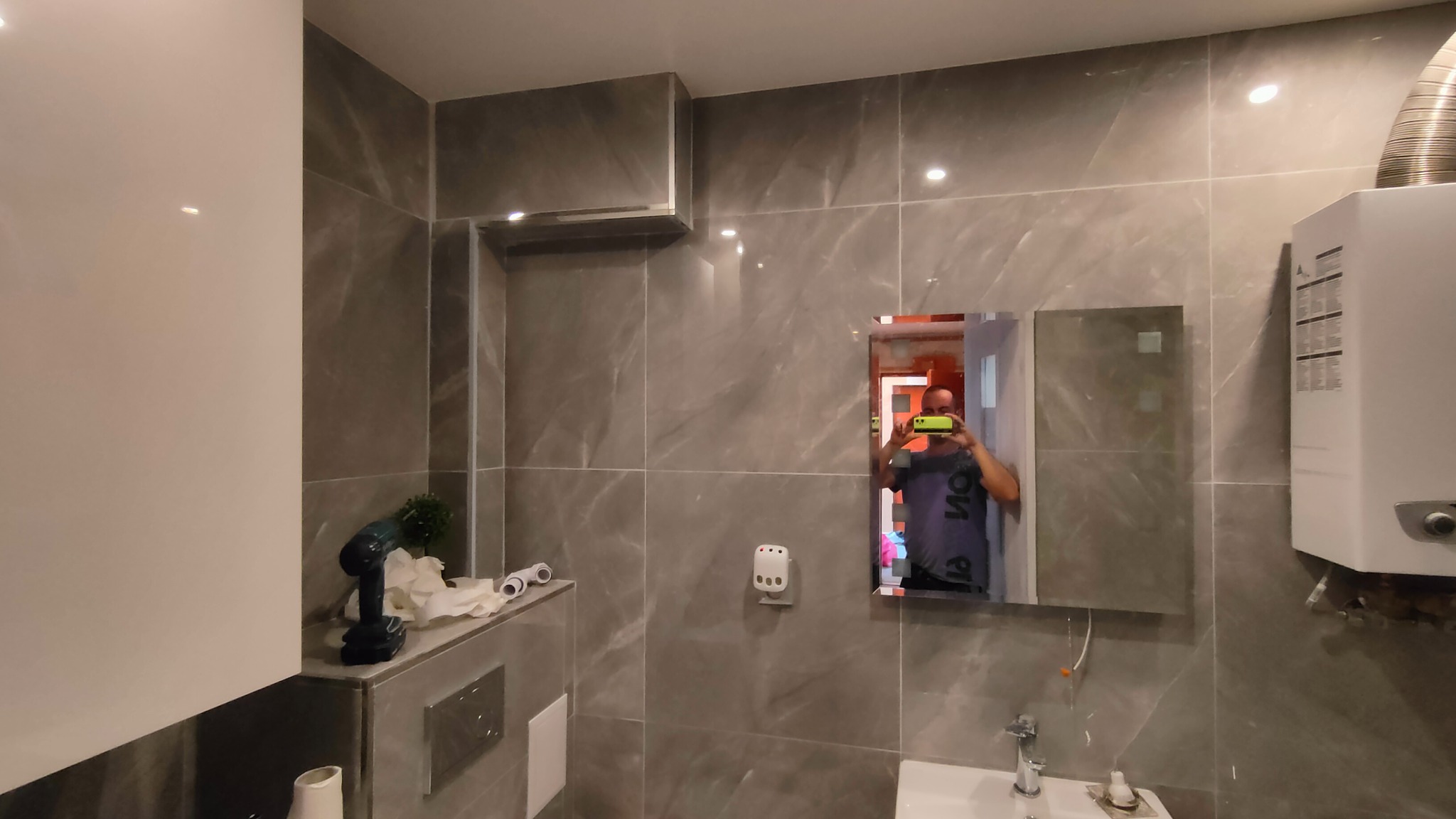 Wykończona łazienka z szarymi płytkami imitującymi marmur, widoczny bojler, lustro z odbiciem osoby robiącej zdjęcie telefonem, umywalka z baterią oraz narzędzia na półce.