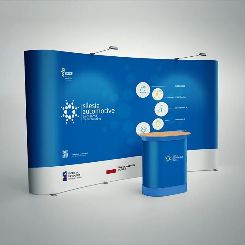 Niebiesko-białe stoisko targowe z logo Silesia Automotive i hasłami promującymi mobilność, cyfryzację, kompetencje, innowacje i kooperację, oświetlone reflektorami, z dodatkowym pulpitem...