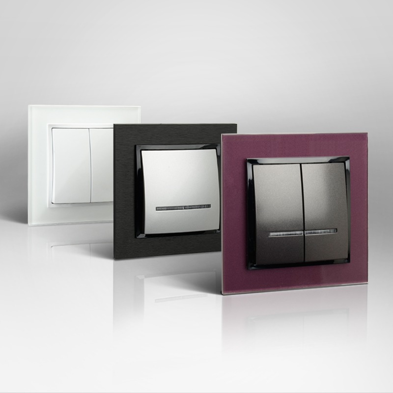 Trzy nowoczesne włączniki światła z różnymi ramkami: białą, czarną i bordową, ustawione w rzędzie na błyszczącej powierzchni, odbijające się w niej.