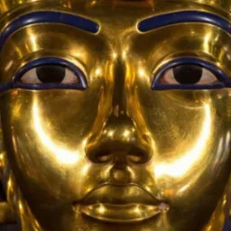 Złota maska pośmiertna Tutanchamona z niebieskimi i turkusowymi detalami, widoczna od frontu, z silnym odbiciem światła na twarzy.