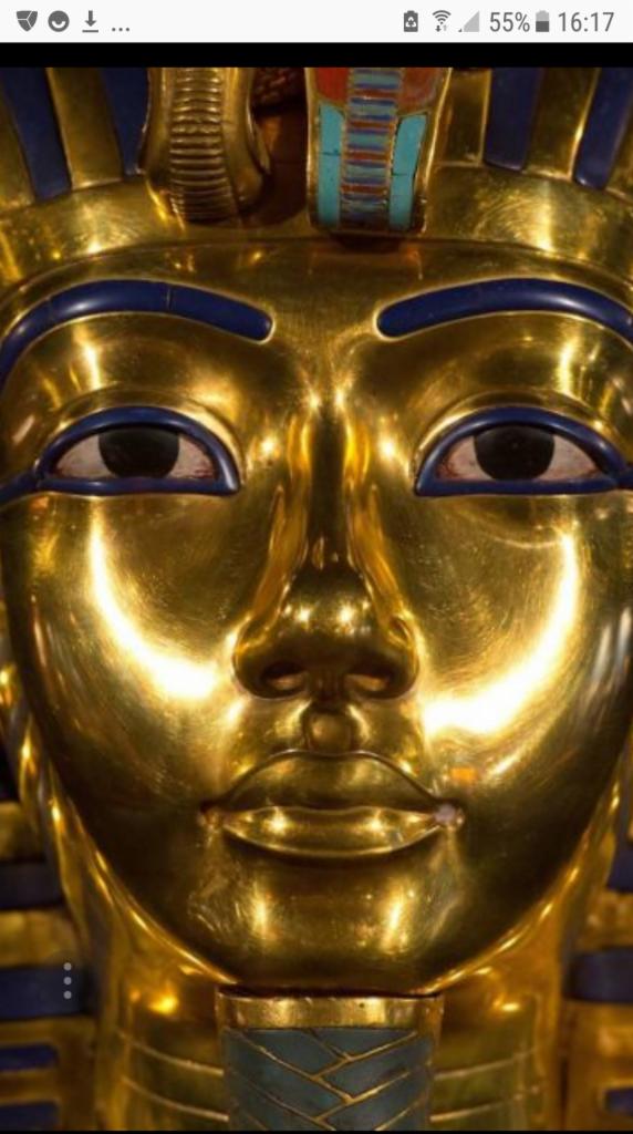 Złota maska pośmiertna Tutanchamona z niebieskimi i turkusowymi detalami, widoczna od frontu, z silnym odbiciem światła na twarzy.