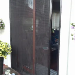 Zewnętrzna moskitiera przesuwna w brązowej ramie, zamontowana na drzwiach tarasowych, widok z zewnątrz, wycieraczka i buty przed wejściem.