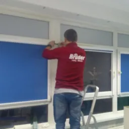 Montaż rolety wewnętrznej w Poznaniu przez instalatora stojącego na drabinie, widoczne okna z zamontowanymi roletami i logo firmy na bluzie montera.