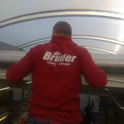 Pracownik w czerwonej bluzie z logo firmy montuje osłony okienne w przeszklonym dachu, widok z dołu na plecy.