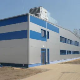 Nowoczesny budynek przemysłowy z szarymi panelami i niebieskimi akcentami, widoczny system wentylacji na dachu, teren wokół budynku częściowo utwardzony.