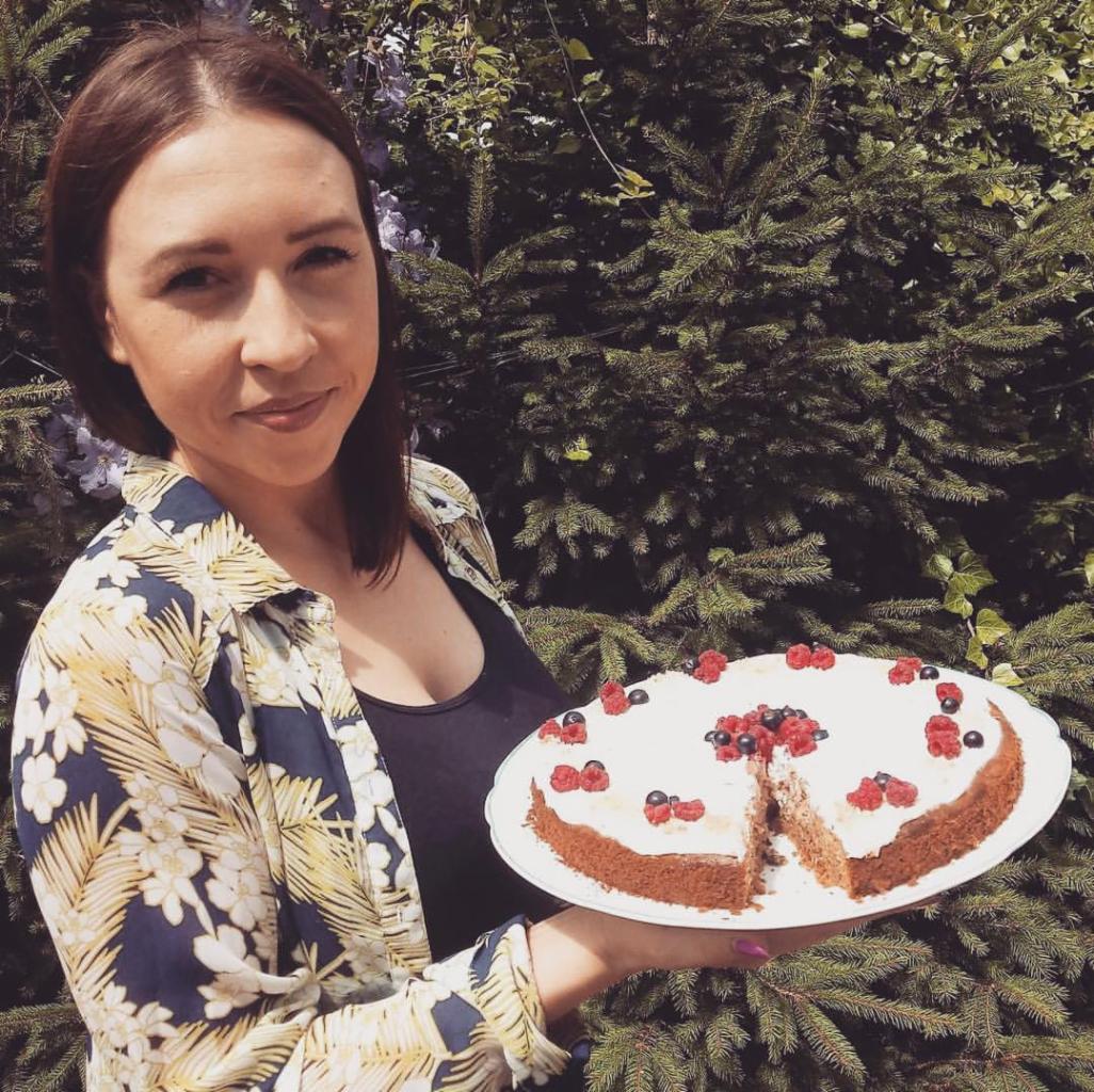 Uśmiechnięta kobieta prezentuje okrągły tort z kremem i owocami leśnymi, z wyciętym kawałkiem, na tle zielonych gałęzi.