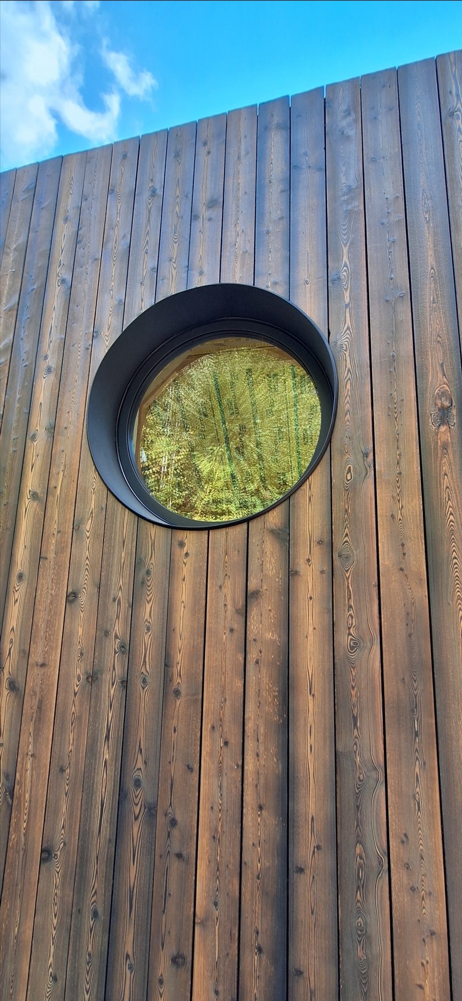 Ściana domu z ciemnego drewna z okrągłym oknem, w którym odbija się las. Nietypowy detal architektoniczny.