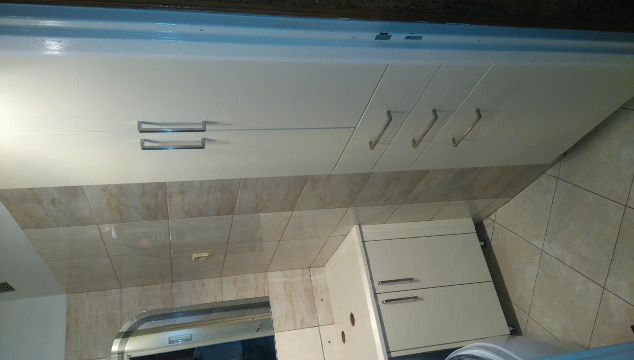 Białe szafki kuchenne z metalowymi uchwytami, beżowe płytki na ścianie i podłodze, widok z dołu z lekkim kątem