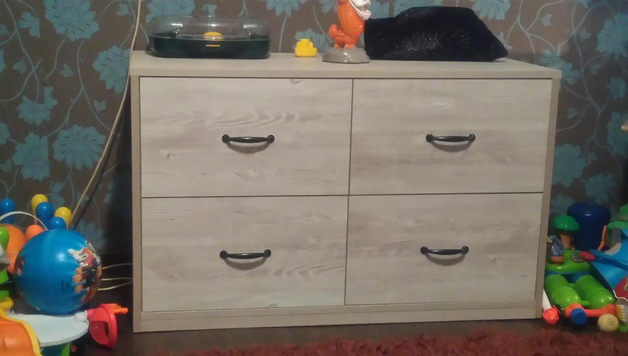 Komoda z czterema szufladami o jasnym, drewnopodobnym wykończeniu, z zabawkami na górze i obok, na tle ściany w kwiaty.