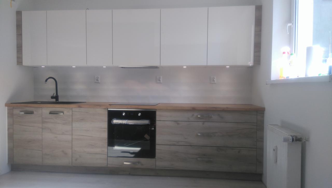 Nowoczesna zabudowa kuchenna z białymi szafkami wiszącymi i drewnopodobnymi szafkami dolnymi, czarny zlew i bateria, okno z widocznymi środkami czystości.