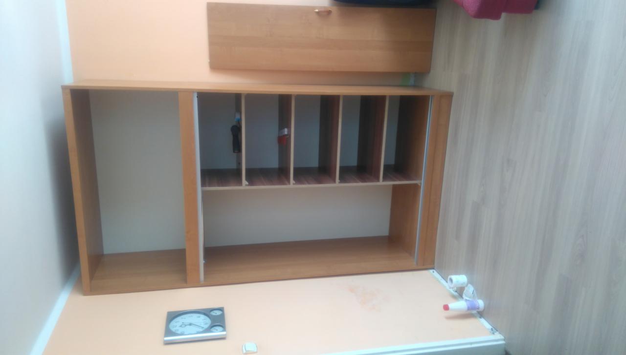 Półka z przegródkami z jasnego drewna, zamontowana na ścianie, z widocznym zegarem i elementami instalacji wodnej w tle