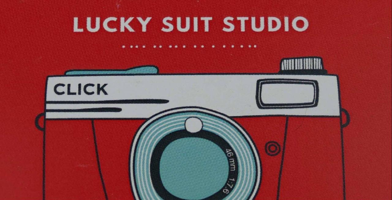 Grafika stylizowanego, czerwonego aparatu fotograficznego z napisem 'CLICK' na białym tle, umieszczona na czerwonym tle z nazwą studia 'LUCKY SUIT STUDIO'.