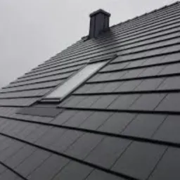 Ciemnoszary dach z dachówki ceramicznej z oknem dachowym i kominem w pochmurny dzień.