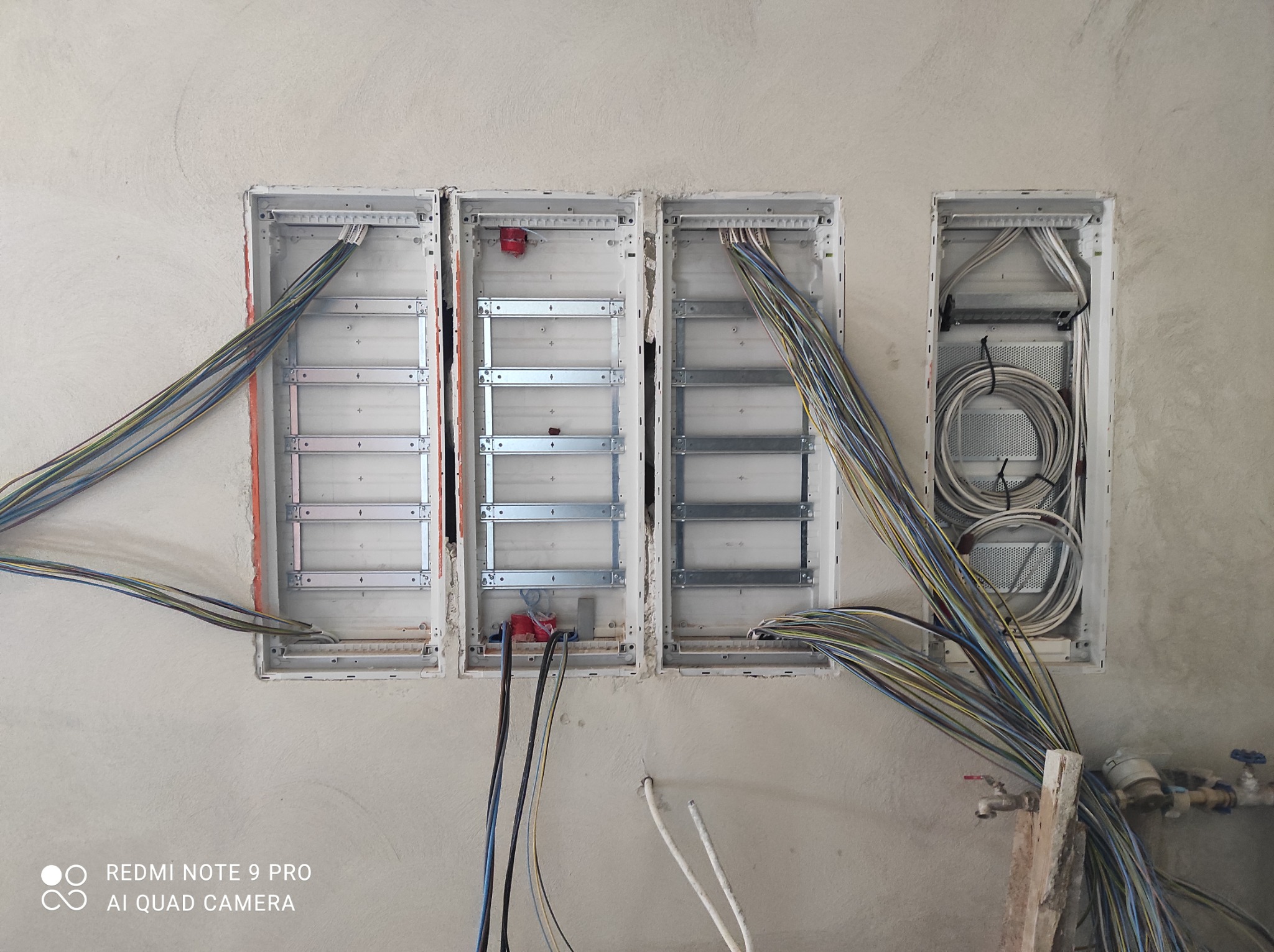 Trzy metalowe skrzynki rozdzielcze w ścianie w trakcie prac montażowych, widoczne kable w różnych kolorach i przekrojach, surowy tynk na ścianie.
