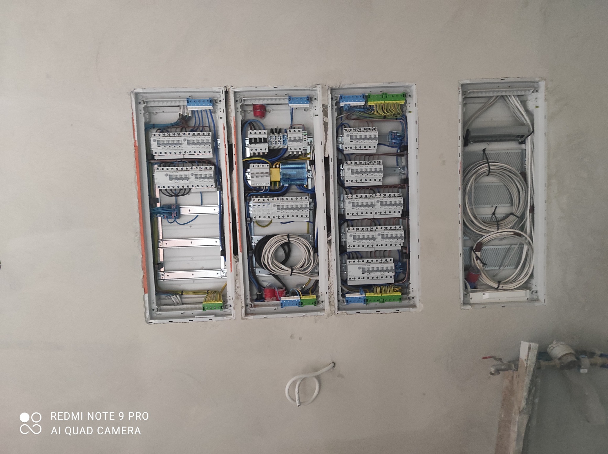 Trzy białe skrzynki elektryczne z zamontowanymi bezpiecznikami i kablami na surowej ścianie w trakcie prac wykończeniowych.